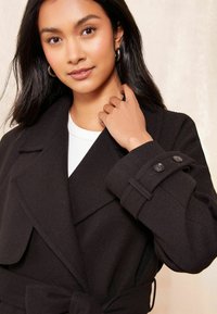 Melnas vilnas maisījuma mētis ar plašu apkakli, dubulto pogu aizdari uz piedurknēm un jostā aizmugurē. Strukturēta auduma, pielāgota dizaina.