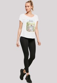 Bílá tričko s krátkým rukávem s ilustrativním designem na přední straně, v kombinaci s černými legínami a černými sportovními botami. Modelka stojí.