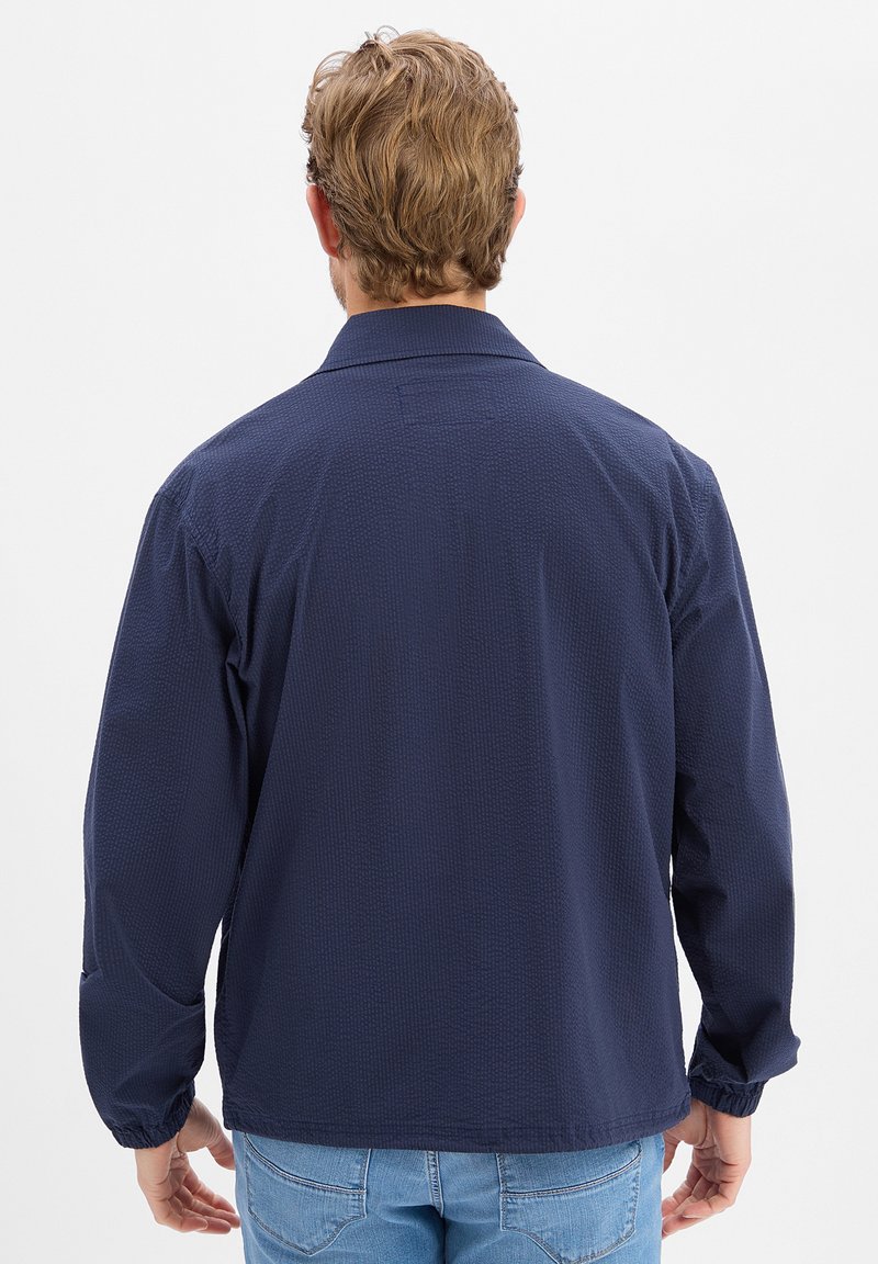 Homme aux cheveux ondulés châtain clair portant une veste texturée bleu marine et un jean bleu clair, vu de dos sur un fond blanc uni.