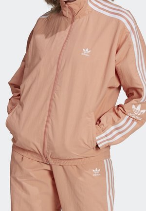 Kobieta w jasnoróżowym dresie Adidas z białymi paskami i logo koniczynki na kurtce i spodniach, ręce w kieszeniach kurtki.