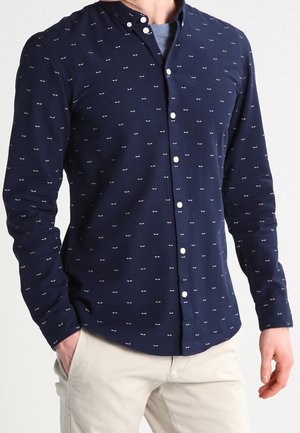Mężczyzna w granatowej koszuli na guziki z małymi białymi wzorami w kształcie oczu i beżowych spodniach, stojący z jedną ręką w kieszeni.