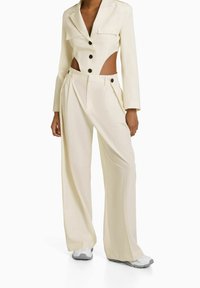 Cream-colored tailored blazer with cut-outs and black buttons paired with wide-legged trousers, featuring a textured finish and subtle tailoring details.