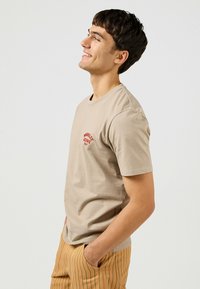 Beige bomullströja med korta ärmar, med en röd broderad logotyp på framsidan. Matches med randiga byxor i tan och grön.