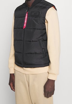 Homme portant un gilet matelassé noir avec fermeture éclair devant, une chemise beige à manches longues et un pantalon beige, debout devant un fond uni.