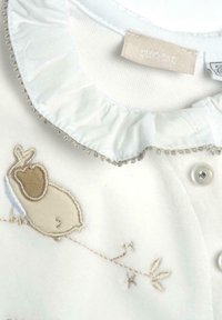 Camisa de tela blanca con un collar con volantes, que presenta un diseño bordado de un pájaro y una rama en color beige. Incluye un cierre de botón.
