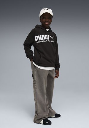 Jeune personne portant un sweat à capuche noir Puma, une casquette blanche, un pantalon ample gris et des chaussons noirs Puma, debout devant un fond dégradé gris.