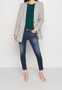 Marynarka w kratę, zielony prążkowany top, ciemne obcisłe jeansy oraz białe sandały na obcasie z paskami. Marynarka ma dopasowany krój. Jeansy posiadają kieszeń na zamek.