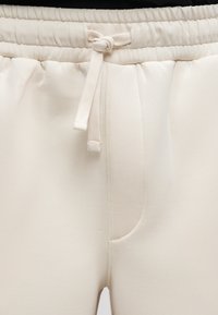 Pantalons de jogging de couleur crème avec une taille élastique, un cordon de serrage et des coutures latérales. Texture lisse avec un design minimaliste.