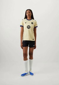 Naine seisab, seljas koorvalge jalgpalli särk Spotify logo ja mustade lühikeste pükste, valgete põlvikute ning siniste tossudega, ühetaolise tausta ees.