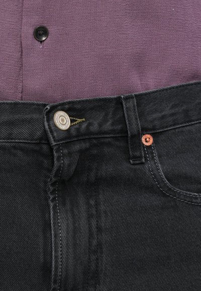 Paarse textuurshirt met zwarte denimjeans met een zilveren knoopsluiting, koperen klinknageldetails en contrasterende stiksels.