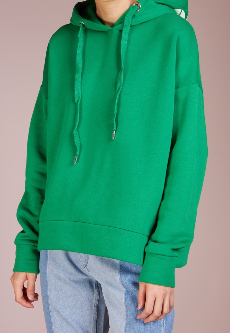 Personne portant un sweat à capuche vert vif avec des cordons et un jean bicolore bleu clair sur un fond uni.