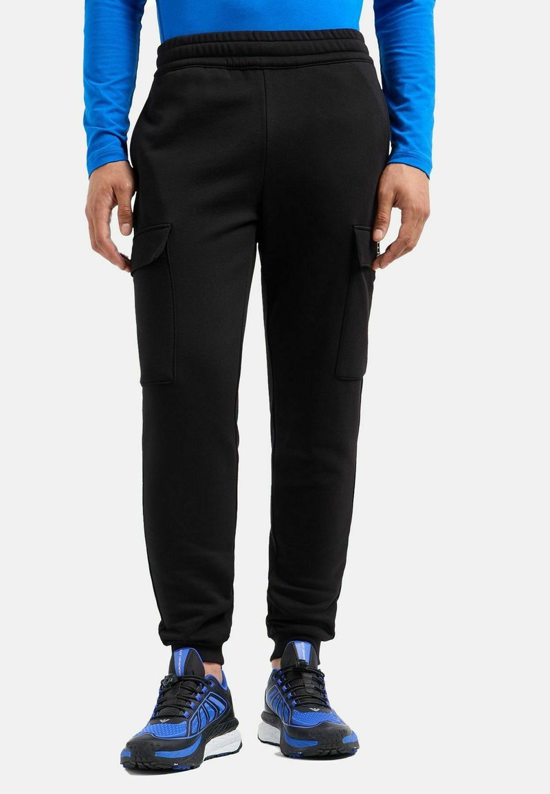 Schwarze Jogginghosen aus einem Baumwollmischgewebe, mit seitlichen Cargo-Taschen, einem elastischen Bund und gerippten Bündchen. Kombiniert mit blauen Sportschuhen.