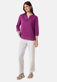Lila Bluse mit langen Ärmeln, V-Ausschnitt und Knopfdetails, kombiniert mit weißen, lässigen Hosen und silbernen Sandalen.