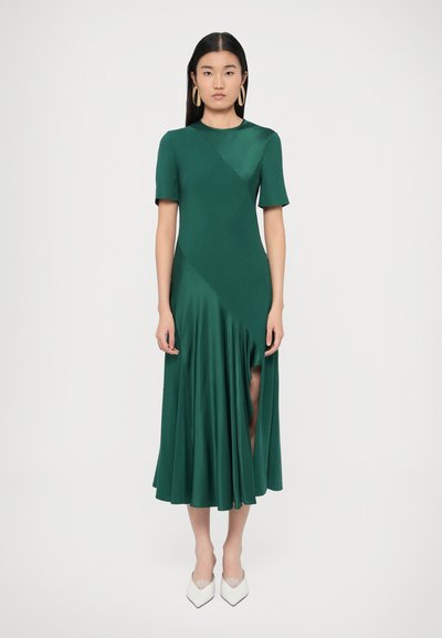 Groene jurk met korte mouwen, een nauwsluitend lijfje en een uitlopende rok. Gladde stof met een diagonale print en een zijsplit. Draagt witte hakken.