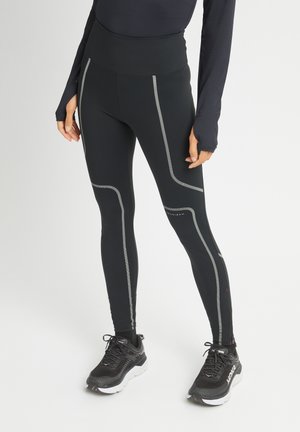 Black high-waisted leggings with reflective grey stitching. Features a smooth texture and a fitted shape, paired with black athletic shoes.