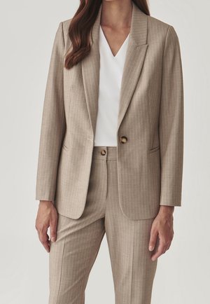 Kobieta ubrana w beżowy garnitur w cienkie prążki z marynarką na jeden guzik i dopasowanymi spodniami na białym topie z dekoltem w serek, stojąca na jednolitym tle.