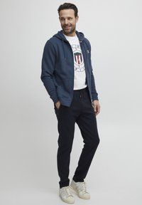 Sweat à capuche navy, t-shirt graphique blanc, joggings sombres et baskets blanches. Le sweat à capuche a des poches avant ; le graphique inclut un design circulaire avec des rayures.