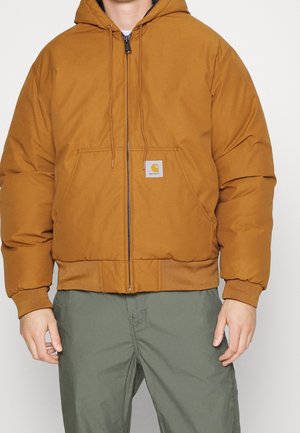 Man draagt een bruine gewatteerde jas met capuchon, een rits aan de voorkant en zakken, gecombineerd met een groene broek, tegen een effen lichte achtergrond.