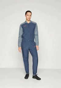 Marinstål och grå Nike-träningsdräkt med en zip-up-jacka och matchande byxor. Jackan har en röd logotyp på bröstet och elastiska muddar.