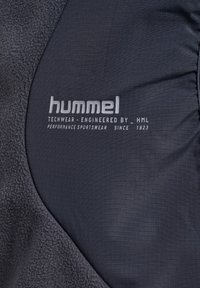 Dunkles Gewebe mit strukturiertem Fleece und Karomuster, versehen mit dem weißen "hummel"-Logo und dem Text "Techwear - Entwickelt von HML Performance Sportswear seit 1923."
