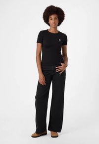 Zwart T-shirt met korte mouwen en een klein logo op de borst, gecombineerd met zwarte wijde broekspijpen en bruine schoenen, in een studiowerkomgeving.