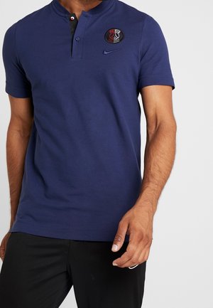 Mężczyzna w niebieskiej koszulce polo z krótkim rękawem z logo Paris Saint-Germain i Nike, w połączeniu z czarnymi spodniami.