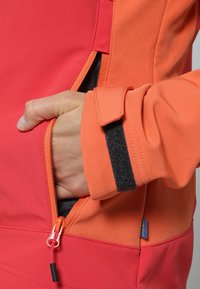 Veste polaire en rouge vif et orange, dotée d'une poche zippée et de poignets ajustables avec une bande velcro. Tissu doux et texturé.