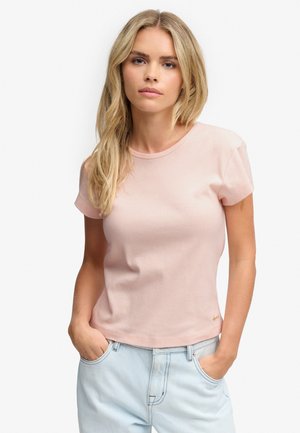 Lyshåret kvinde med bølget hår iført en lyserød kortærmet t-shirt og lyseblå jeans med hænderne i lommerne.