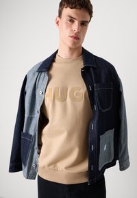 Veste en denim avec des manches contrastantes bleu et bleu clair, associée à un sweat-beige avec des lettres en relief "HUGO".