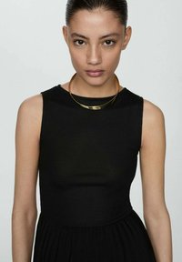 Schwarzes ärmelloses Kleid mit geripptem Muster und einer Länge bis unter das Knie. Ergänzt durch eine goldene metallische Halskette.