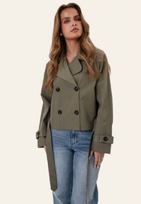 Kvinde iført en kort olivengrøn dobbeltradet jakke med knapmanchetter og blå jeans, stående mod en ensfarvet baggrund.
