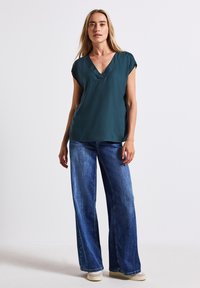 Maglietta verde acqua a V in tessuto morbido con maniche corte, abbinata a jeans a gamba larga in lavaggio chiaro e scarpe chiare. Design semplice e casual.