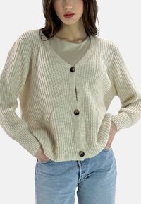 Kerniger cremefarbener Cardigan mit V-Ausschnitt, großen dunklen Knöpfen und gerippter Struktur. Kombiniert mit einem hellen T-Shirt und blauen Jeans.