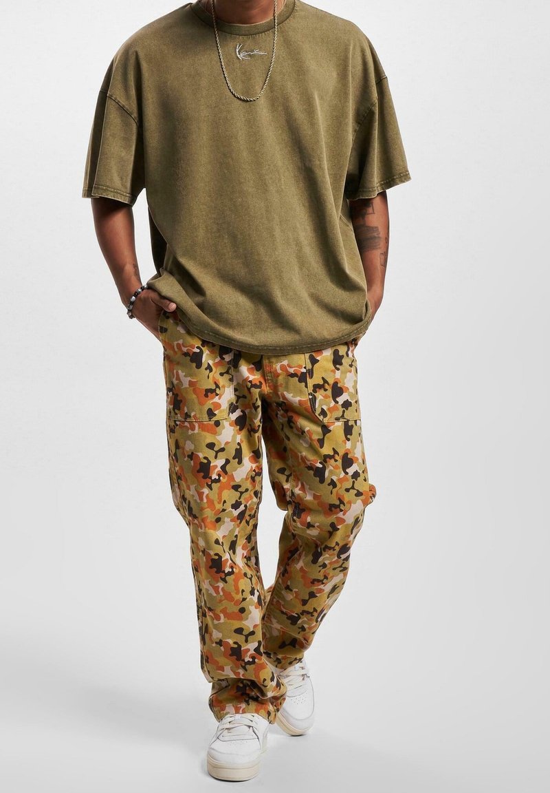 Olivově zelené oversized tričko s krátkým rukávem, doplněné kalhotami s maskáčovým vzorem v oranžové, hnědé a béžové barvě. Bílé tenisky dotvářejí celý outfit.