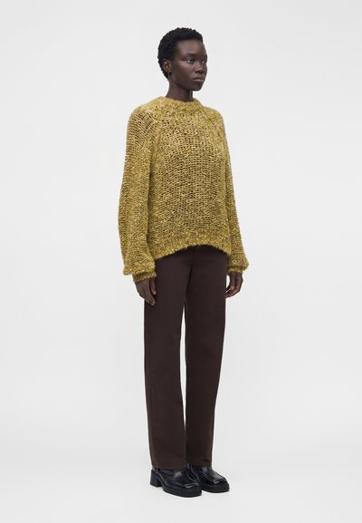 Femme portant un pull en maille jaune ample, un pantalon droit marron foncé et des chaussures à plateforme noires, debout sur un fond blanc uni.