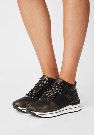 Jambes portant des baskets à lacets noires et bronze avec des semelles blanches, debout sur un fond gris clair uni.