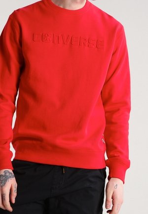 Personne portant un sweat-shirt rouge avec le logo "Converse" en relief sur la poitrine et un pantalon noir, montrant des mains tatouées de chaque côté.