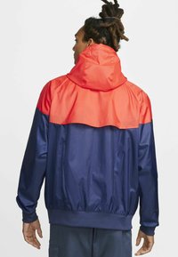 Jūros mėlynasis ir raudonas vėjo nepraleidžiantis švarkas, su gobtuvu, elastinėmis rankovėmis ir spalvų blokų dizainu, pagamintas iš lygaus, blizgaus audinio.