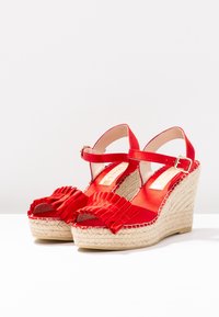 Sandalias de cuña rojas con parte superior de tela, que cuentan con acentos de volantes y suela de esparto. Correa ajustable en el tobillo con hebilla de metal.