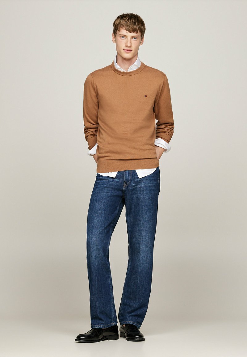 Brauner Strickpullover mit Rundhalsausschnitt, darunter ein weißes Hemd mit Kragen, kombiniert mit blauen geraden Jeans und schwarzen Schuhen.