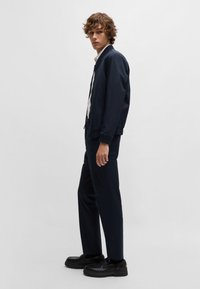Blouson de pilote marine avec col côtelé, porté sur une chemise blanche. Pantalon sombre slim-fit et chaussures noires épaisses. Design simple et minimaliste.