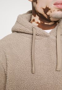 Beige fleecová mikina s texturovaným povrchem, vybavená šňůrkou na kapuci a volným střihem. Tkanina vypadá měkce a teple.