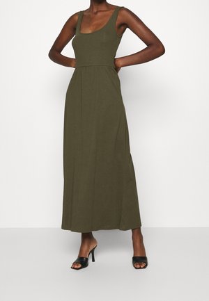 Femme portant une robe maxi sans manches vert olive avec un décolleté en U et des talons ouverts noirs, debout les mains sur les hanches.