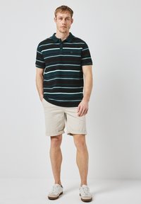 Svart och turkosrandig pikétröja med krage, kombinerad med uppvikta beige shorts och vita sneakers. Tröjan har en liten logodetalj.