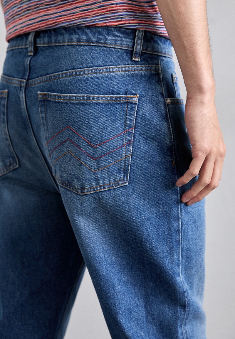 Blå denimjeans med en bakficka som har en sicksackssömnad i röd, orange och gul. Tyget har en klassisk struktur och passform.