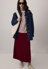 Žena v tmavomodrej oversized denovej bunde, červeno-bielom prúžkovanom tričku, dlhých červených sukňa, bielych teniskách, slnečných okuliaroch a s béžovou chlpatou kabelkou.