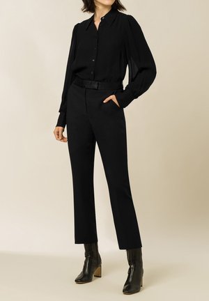Vrouw draagt een zwarte blouse met knopen en opgepofte mouwen, zwarte hooggetailleerde broek en zwarte enkellaarzen met blokhakken.