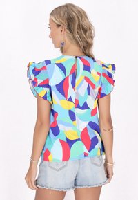Mehrfarbige Bluse mit kurzen Rüschensäumen, die abstrakte Formen in Blau, Rot, Gelb und Grün zeigt. Knopfverschluss auf der Rückseite.
