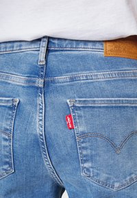 Jeans de mezclilla azul claro con una etiqueta de cuero marrón en la cinturilla y etiqueta de logo roja. Presenta bolsillos traseros cosidos con un diseño clásico.