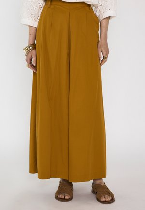 Vrouw draagt mosterdkleurige wijde broek, witte blouse met opengewerkte details, bruine sandalen en gouden armbanden aan de linkerpols en ringen aan de vingers.